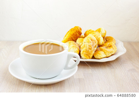 Milk coffee cup with saucer and a croissants on light wooden background close up. Breakfast concept. Milk coffee cup with saucer and a croissants on light wooden background close up. Breakfast concept. 64888580