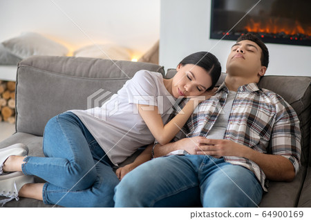 Dark-haired female resting her head on brunette male shoulder, both resting on sofa Dark-haired female resting her head on brunette male shoulder, both resting on sofa 64900169