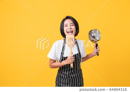 Happy young asian woman wear on an apron and hands showing spatula with pot Happy young asian woman wear on an apron and hands showing spatula with pot 64904316