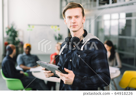 Half-length portrait of cheerful young manager or designer dressed in casual outfit and earphones, having work break in office, holding digital tablet. successful designers team having productive job 64918226