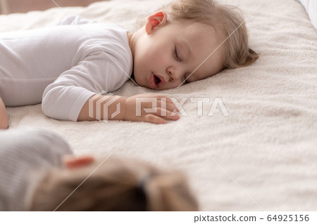 Childhood, sleep, relaxation, family, lifestyle concept - two young children 2 and 3 years old dressed in white and beige bodysuit sleep on a beige and white bed at lunch holding hands top view. Childhood, sleep, relaxation, family, lifestyle concept - two young children 2 and 3 years old dressed in white and beige bodysuit sleep on a beige and white bed at lunch holding hands top view. 64925156