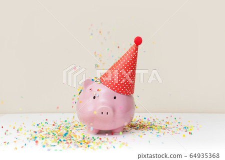 Pink piggy bank with birthday cap and color confetti on beige background Pink piggy bank with birthday cap and color confetti on beige background 64935368