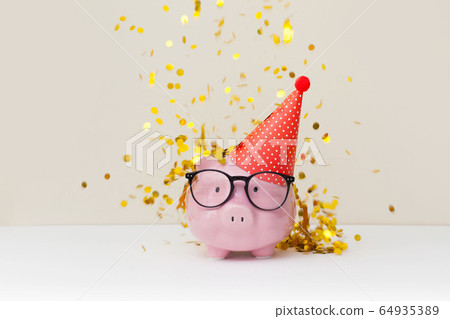 Pink piggy bank with birthday cap on beige background Pink piggy bank with birthday cap on beige background 64935389