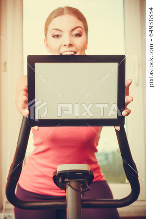 Woman on exercise bike holding pc tablet copyspace 64938334