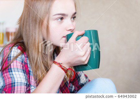 Attractive blonde girl is drinking tea from a large green mug in the kitchen. The concept of spending time during self-isolation of a virus pandemic Attractive blonde girl is drinking tea from a large green mug in the kitchen. The concept of spending time during self-isolation of a virus pandemic 64943002