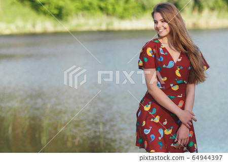 Young attractive girl in a burgundy dress by the river. Sunset Young attractive girl in a burgundy dress by the river. Sunset 64944397