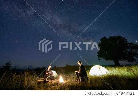 Friends travellers spending time in the mountains sitting around bonfire in camping under beautiful starry sky 64947421