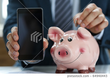 In hands smartphone, man puts coin in piggy bank 64948443
