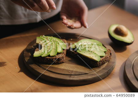 Woman making vegan sandwich using rye toasted bread avocado and sesame seeds. Woman sprinkle with sesame seeds on sandwich 64950089