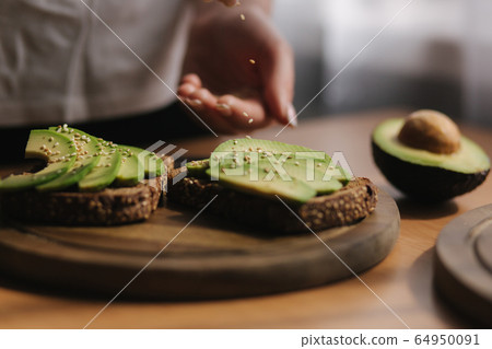 Woman making vegan sandwich using rye toasted bread avocado and sesame seeds. Woman sprinkle with sesame seeds on sandwich Woman making vegan sandwich using rye toasted bread avocado and sesame seeds. Woman sprinkle with sesame seeds on sandwich 64950091