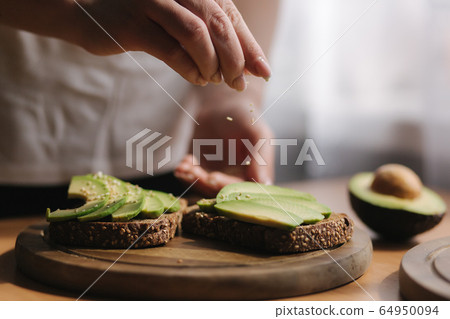 Woman making vegan sandwich using rye toasted bread avocado and sesame seeds. Woman sprinkle with sesame seeds on sandwich Woman making vegan sandwich using rye toasted bread avocado and sesame seeds. Woman sprinkle with sesame seeds on sandwich 64950094