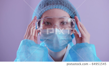 Asian woman doctor in a mask and a protective suit puts on safety glasses and looks at the camera. Coronavirus epidemic, concept. Asian woman doctor in a mask and a protective suit puts on safety glasses and looks at the camera. Coronavirus epidemic, concept. 64951545
