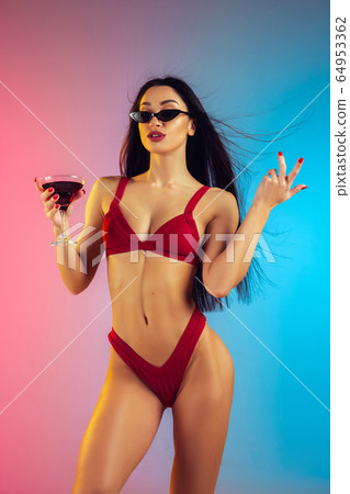 Fashion portrait of young fit and sportive woman with cocktail in stylish red luxury swimwear on gradient background. Perfect body ready for summertime. 64953362
