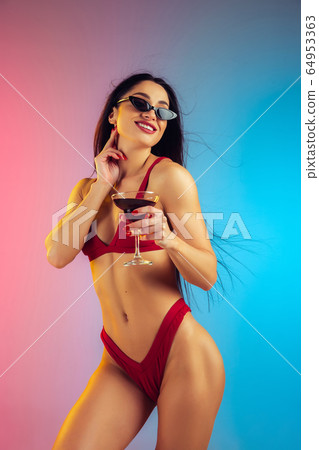 Fashion portrait of young fit and sportive woman with cocktail in stylish red luxury swimwear on gradient background. Perfect body ready for summertime. 64953363