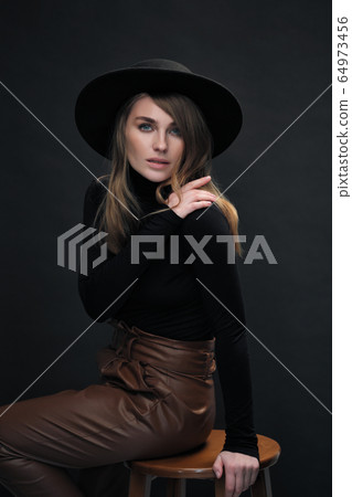 Portrait of young beautiful blond woman with light makeup and blue eyes touching her face. Dark background. Black tight sweater and hat with fields. 64973456