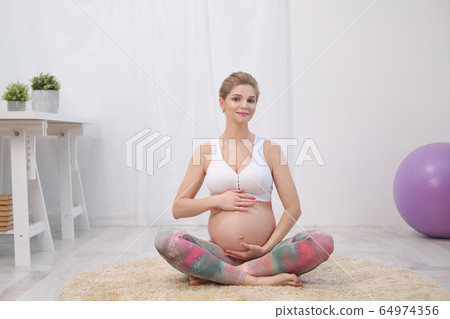 A young beautiful pregnant Caucasian woman sits on the soft carpet of the house. He is engaged in fitness and doing exercises. Sportswear. 64974356