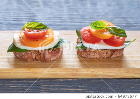 Two bruschetta Caprese: toasted bread slices, red and yellow tomatoes, mozzarella cheese, green Basil, herbs, olive oil on wooden plank on grey wooden table. Two bruschetta Caprese: toasted bread slices, red and yellow tomatoes, mozzarella cheese, green Basil, herbs, olive oil on wooden plank on grey wooden table. 64975677