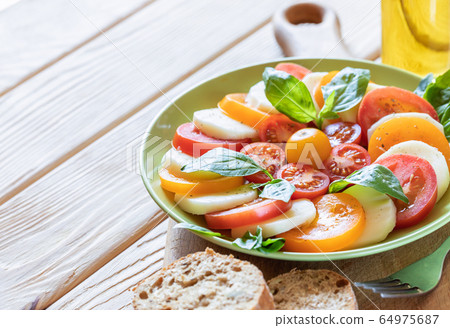 Close-up green plate with traditional Italian Caprese salad: red and yellow tomatoes, mozzarella cheese, Basil, herbs, olive oil on kitchen board on wooden table with copy space. Selective focus. Close-up green plate with traditional Italian Caprese salad: red and yellow tomatoes, mozzarella cheese, Basil, herbs, olive oil on kitchen board on wooden table with copy space. Selective focus. 64975687