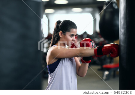 Athletic young brunette woman in sportswear and red boxing gloves trains bumps on a punching bag in a fitness gym. 64982368