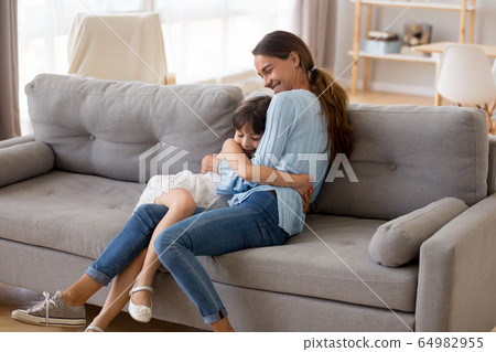 Happy mother hugging cute little girl sitting together on sofa Happy mother hugging cute little girl sitting together on sofa 64982955