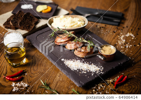 Pork tenderloin with mashed potatos, bacon and mustard sauce covered with a branch of smoking rosemary on black board. Delicious healthy traditional grilled meat food closeup served on a table for Pork tenderloin with mashed potatos, bacon and mustard sauce covered with a branch of smoking rosemary on black board. Delicious healthy traditional grilled meat food closeup served on a table for 64986414
