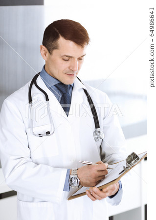 Doctor man using clipboard for filling up medication history records. Perfect medical service in clinic. Physician at work in hospital. Medicine and healthcare concepts Doctor man using clipboard for filling up medication history records. Perfect medical service in clinic. Physician at work in hospital. Medicine and healthcare concepts 64986641
