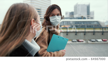 Side view two young Caucasian doctor women wearing face masks, walking on empty city street with clipboards on lockdown. Side view two young Caucasian doctor women wearing face masks, walking on empty city street with clipboards on lockdown. 64987000