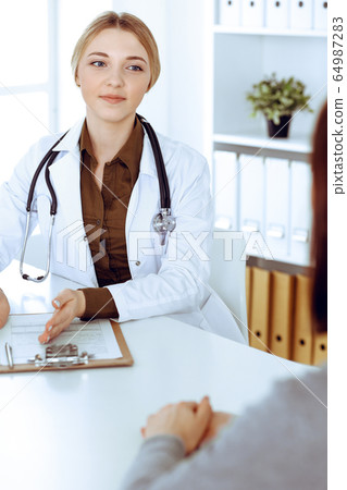 Young woman doctor and patient at medical examination in hospital office. Khaki colored blouse of therapist looks good. Medicine, healthcare and doctor's appointment concept Young woman doctor and patient at medical examination in hospital office. Khaki colored blouse of therapist looks good. Medicine, healthcare and doctor's appointment concept 64987283