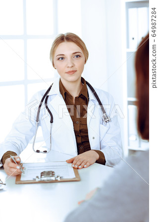 Young woman doctor and patient at medical examination in hospital office. Khaki colored blouse of therapist looks good. Medicine, healthcare and doctor's appointment concept Young woman doctor and patient at medical examination in hospital office. Khaki colored blouse of therapist looks good. Medicine, healthcare and doctor's appointment concept 64987284