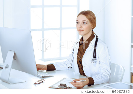 Young woman doctor at work in hospital looking at desktop pc monitor. Physician controls medication history records and exam results. Medicine and healthcare concept Young woman doctor at work in hospital looking at desktop pc monitor. Physician controls medication history records and exam results. Medicine and healthcare concept 64987285