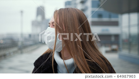 Epidemic quarantine. Portrait of young Caucasian woman wearing mask outdoors, turning head to camera in empty street. Epidemic quarantine. Portrait of young Caucasian woman wearing mask outdoors, turning head to camera in empty street. 64987903