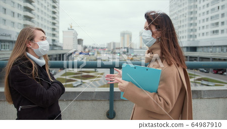 Epidemic control social care workers. Two young Caucasian female social care employees in face masks talk outside. 64987910