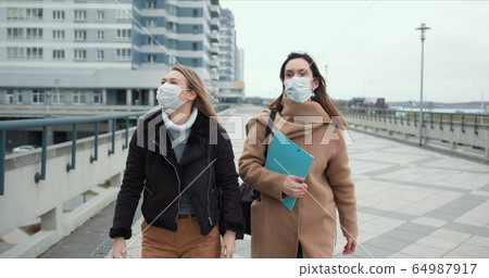 Two young doctor women wear masks walking in empty windy city street. Fear and danger atmosphere on epidemic quarantine. Two young doctor women wear masks walking in empty windy city street. Fear and danger atmosphere on epidemic quarantine. 64987917