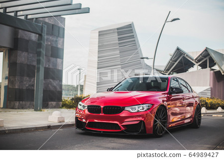 Red model sedan on the empty city roads, across modern buildings, front view Red model sedan on the empty city roads, across modern buildings, front view 64989427