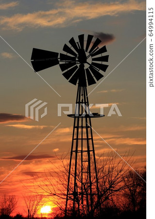 Kansas colorful Sunset with a Windmill silhouette with clouds and tree's and the Sun. 64994115