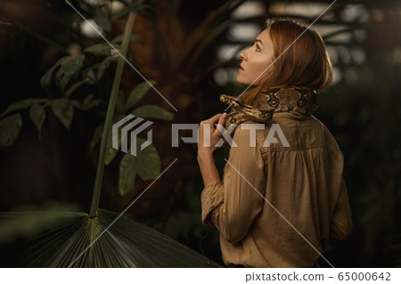 A beautiful girl with natural make-up and red hair stands in the jungle among exotic plants with snake. 65000642