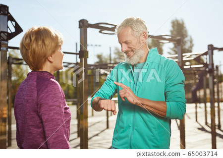 Checking fitness results. Happy senior man in sportswear looking at smartwatch and smiling while exercising with his wife at outdoor gym. Active family couple Checking fitness results. Happy senior man in sportswear looking at smartwatch and smiling while exercising with his wife at outdoor gym. Active family couple 65003714