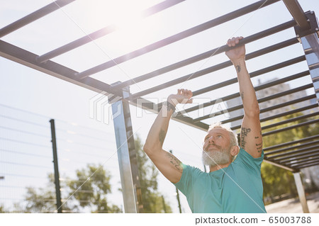 Strong muscles. Senior man with tattoos on his arms climbing monkey bars while working out at outdoor gym. Sport, workout 65003788