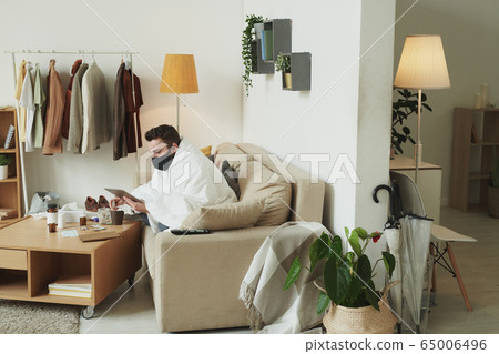 Sick young man in black mask using touchpad while working remotely Sick young man in black mask using touchpad while working remotely 65006496