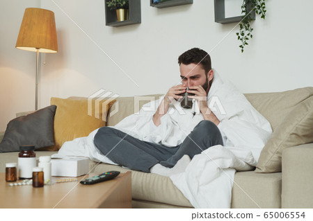 Young man with coronavirus or flu drinking hot tea or water on couch Young man with coronavirus or flu drinking hot tea or water on couch 65006554