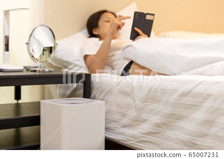 Air purifier with woman using cell phone relaxing in bedroom for cleaning and removing dust PM2.5 HEPA in home. Air purifier with woman using cell phone relaxing in bedroom for cleaning and removing dust PM2.5 HEPA in home. 65007231