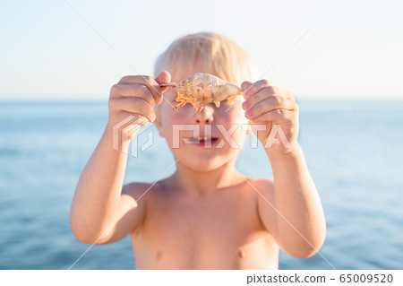 Cute blond boy holding a shell on sea background. Cute blond boy holding a shell on sea background. 65009520