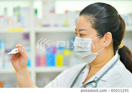 Pharmacist wearing N95 mask with thermometer and stethoscope on many medicine shelf background. Coronavirus (COVID-19)concept of disease, flu treatment and protection Pharmacist wearing N95 mask with thermometer and stethoscope on many medicine shelf background. Coronavirus (COVID-19)concept of disease, flu treatment and protection 65011291