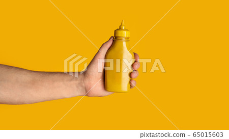 Closeup of millennial guy showing squeeze bottle of mustard on orange background, mockup for design. Panorama Closeup of millennial guy showing squeeze bottle of mustard on orange background, mockup for design. Panorama 65015603