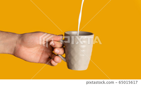 Unrecognizable young man holding cup of hot drink with milk pouring into it, orange background. Panorama Unrecognizable young man holding cup of hot drink with milk pouring into it, orange background. Panorama 65015617
