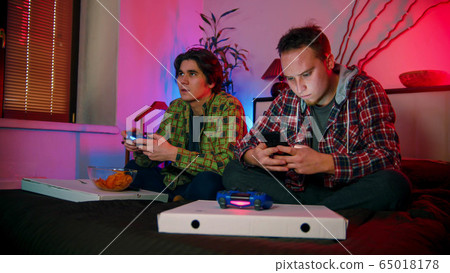 Two young men sitting on the bed - one of them playing the game and other sitting in his phone 65018178
