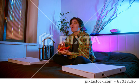 Young interested man sitting on the bed watching TV and eating chips 65018180