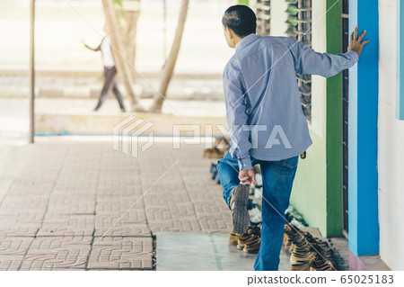 Back view of male teacher stand with his right hand against the wall to easily wear shoes in front of classroom. 65025183