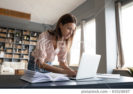 Smiling woman standing near desk, using laptop at home 65028697