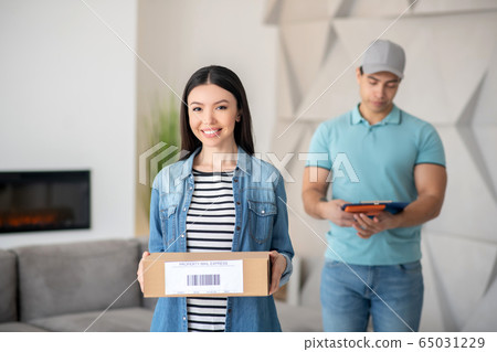 Dark-haired female holding brown cardboard box, brunette male courier standing behind her with clip folder Dark-haired female holding brown cardboard box, brunette male courier standing behind her with clip folder 65031229
